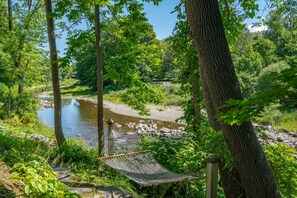 A hammock in a forested area with a river and a bench in the background. - Historic Riverside Home with Sauna & Covered Porch (Woodstock)