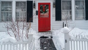 A white building with a red door, black shutters, and a small Christmas wreath. - Historic Riverside Home with Sauna & Covered Porch (Woodstock)
