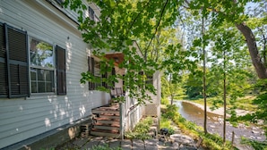 A white house with a porch, steps leading to a wooden deck, and a river in the background. - Historic Riverside Home with Sauna & Covered Porch (Woodstock)