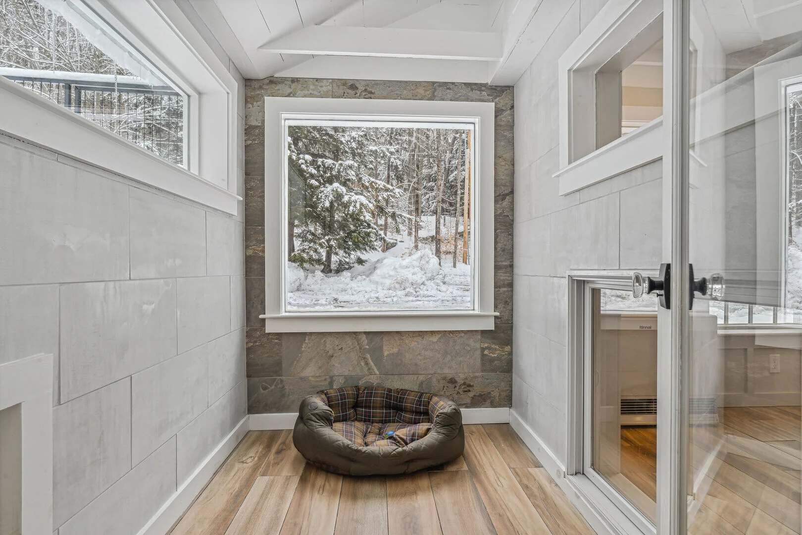 A corner room with a stone wall, a wooden floor, a pet bed, and a large window with a snowy forest view.