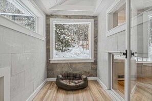 A corner room with a stone wall, a wooden floor, a pet bed, and a large window with a snowy forest view.