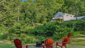 Une maison dotée d’une terrasse, entourée d’une forêt dense, et trois chaises Adirondack disposées face à un foyer extérieur.