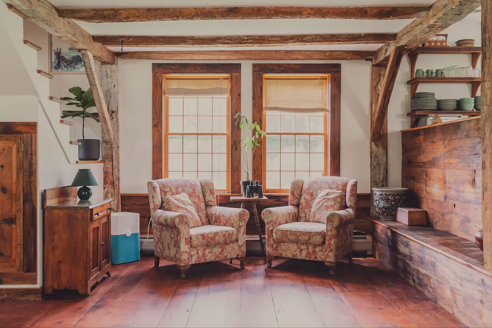 Une pièce rustique avec des poutres en bois, deux fenêtres, une table avec des chaises et des étagères remplies de vaisselle.