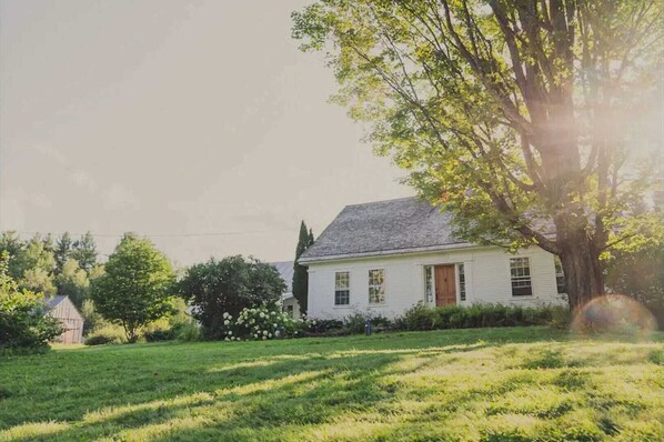 A white house with a sloped roof, surrounded by a grassy yard and trees. - Historic Farmhouse on Working VT Farm & Hot Tub (Woodstock)