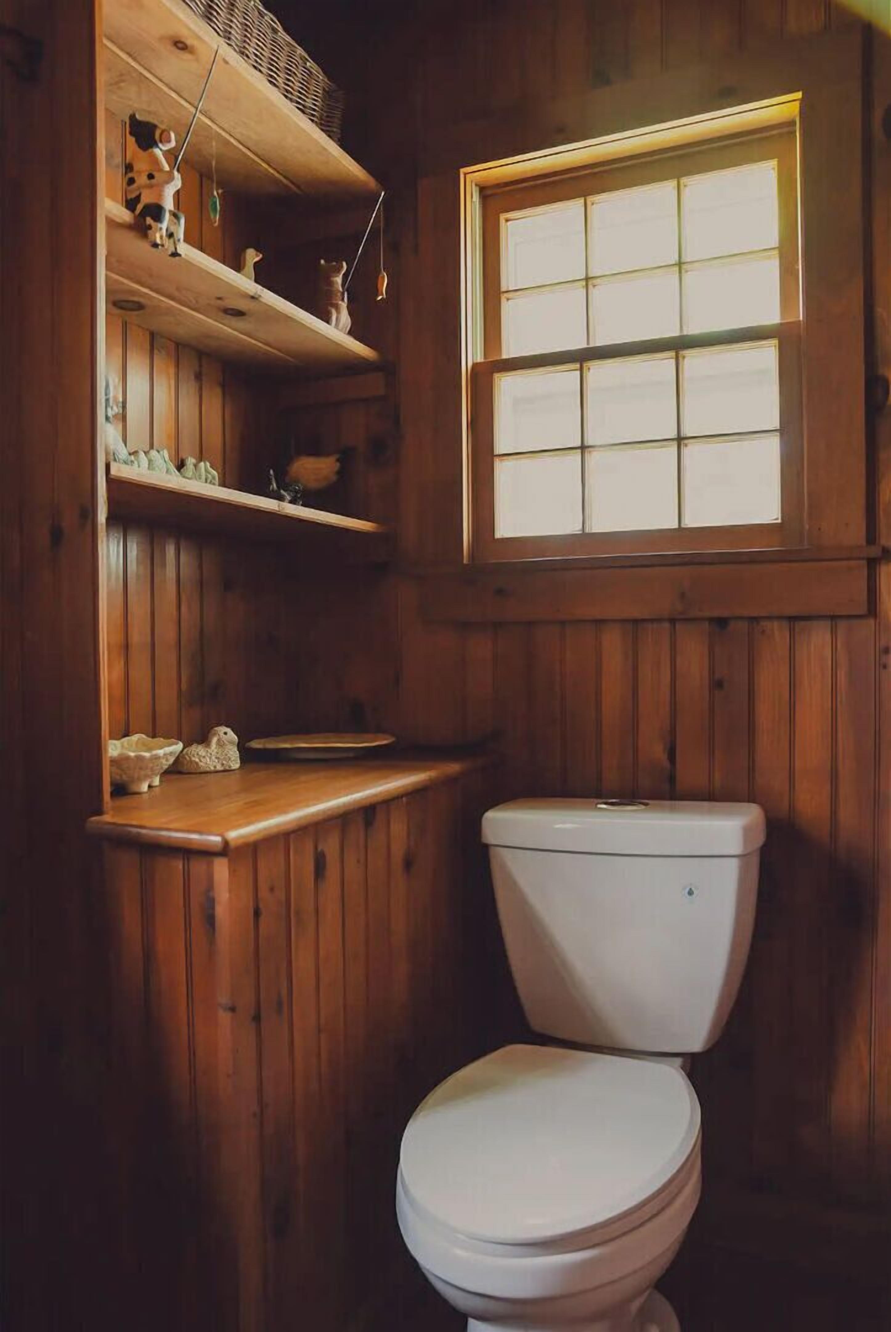 Une salle de bain en bois, avec des toilettes, des étagères ornées d’objets décoratifs et une fenêtre.