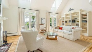 A spacious living room with a white sofa, a glass coffee table, and a white armchair. There is a fireplace, a bookshelf, and a large window with curtains. - Norwich Estate with Pond, Pool & Mountain Views (Norwich)