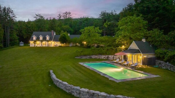 A large, well-maintained lawn with a swimming pool surrounded by stone walls, multiple houses with lit windows, and a gazebo. - Norwich Estate with Pond, Pool & Mountain Views (Norwich)