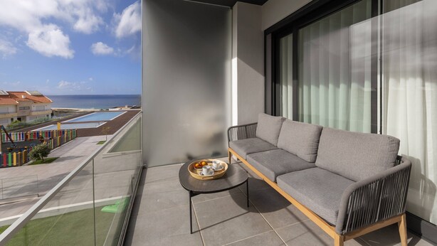Un balcón con un sofá gris, una mesita redonda con una bandeja de frutas y tazas, y vistas a una piscina y edificios.