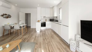 Una cocina moderna con armarios blancos, una zona de comedor con sillas grises y un televisor a la derecha.