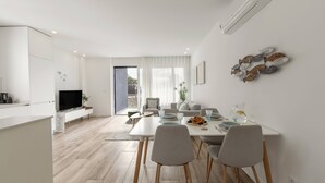 Una cocina moderna con muebles blancos, un televisor de pantalla plana y una zona de comedor con una mesa blanca y sillas grises.