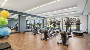 A modern gym with various cardio and strength training equipment, large windows, and a wooden floor.