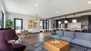 A modern living room with a gray sectional sofa, wooden coffee table, and purple armchairs. A dining area with wooden chairs and a table is visible. The kitchen area features dark cabinets and a microwave.