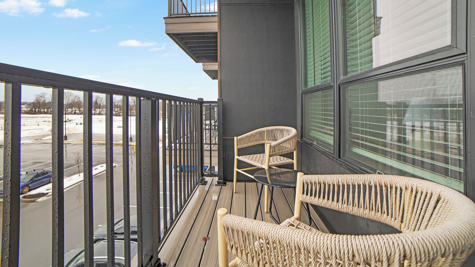A balcony with a wicker chair and a table, overlooking a parking area.
