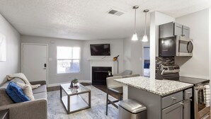 A modern kitchen with a built-in microwave, oven, and granite countertop. There is a dining area with a table and chairs, and a living room with a couch and a fireplace. - Landing Grand Prairie (Grand Prairie)
