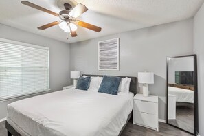 A bedroom with a bed, bedside lamps, a ceiling fan, a window with blinds, a wall-mounted artwork, and a mirror. - Landing Grand Prairie (Grand Prairie)