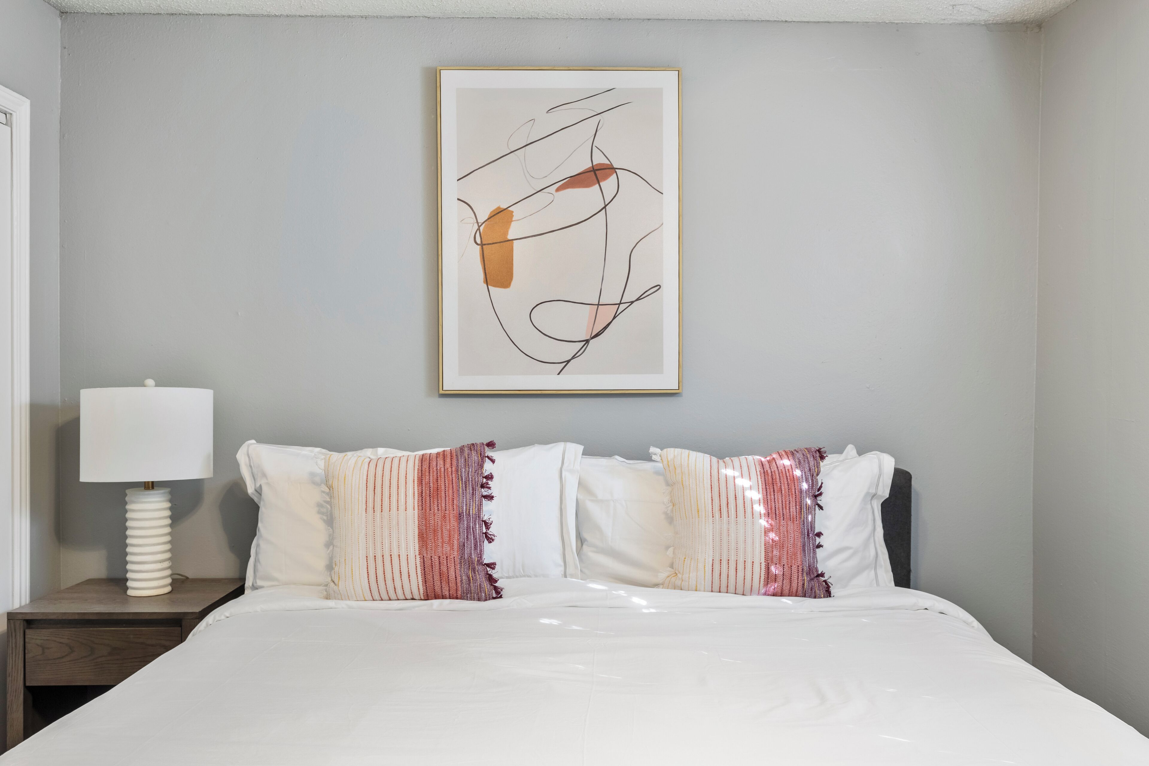 Una cama bien hecha con ropa de cama blanca y almohadas a rayas, una mesita de noche con lámpara, y una obra de arte abstracta enmarcada en la pared.