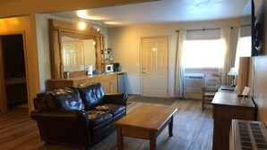 Ein Wohnzimmer mit Ledersofa, Couchtisch aus Holz und einer Küchenzeile mit Mikrowelle und Kaffeemaschine.