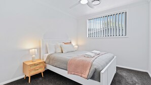 A modern bedroom with a white bed, a nightstand, a window with blinds, and a ceiling fan.