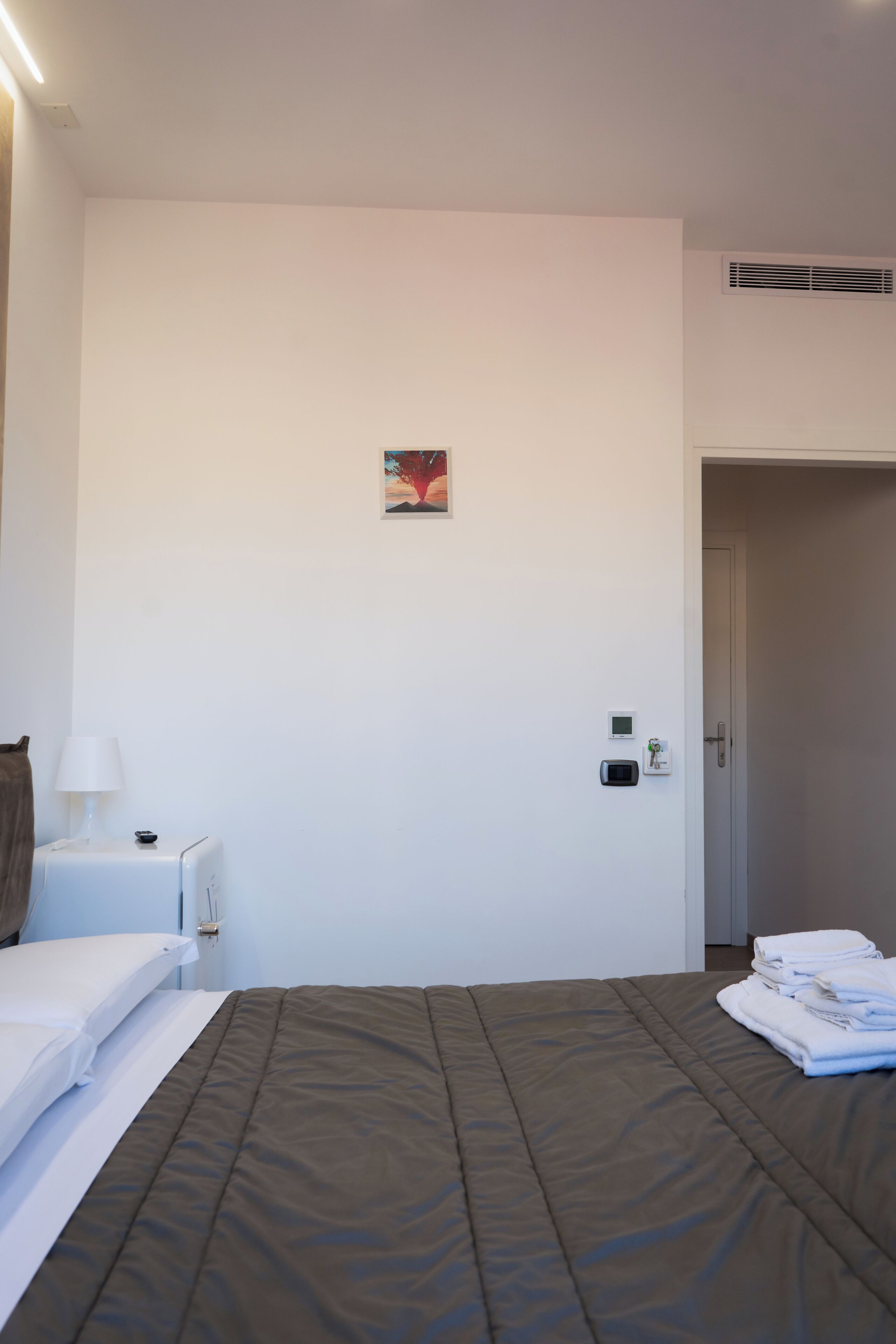 Ein Hotelzimmer mit einem Bett, einem Nachttisch mit Lampe, einem kleinen Bild an der Wand, einer Tür und einer Wand-Klimaanlage.