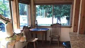 Une pièce avec un plancher en bois, une table ronde entourée de chaises, un comptoir de bar et une grande fenêtre donnant sur des arbres et une voiture garée à l’extérieur.
