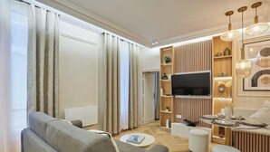 Ein modernes Wohnzimmer mit Sofa, Hocker, Fernseher und einem Essbereich mit Tisch für zwei Personen.