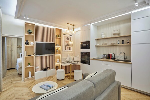 Ein modernes Wohnzimmer mit grauem Sofa, Flachbildfernseher, Holzregalen und einer Küchenzeile mit Mikrowelle und Toaster.