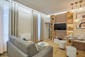 Ein modernes Wohnzimmer mit einer Couch, einem Hocker, einem Fernseher und einem Essbereich mit Tisch für zwei Personen.