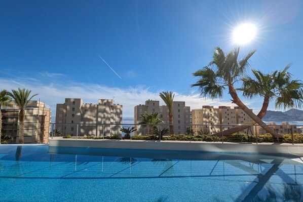 A swimming pool with clear blue water, surrounded by palm trees and buildings with a view of the sea.