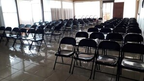 Un salón de conferencias con filas de sillas negras y mesas, ventanas amplias y piso de baldosas.