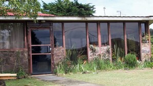 Una casa de un piso con puerta y ventanas de vidrio, rodeada de un jardín con diversas plantas y un muro de piedra.
