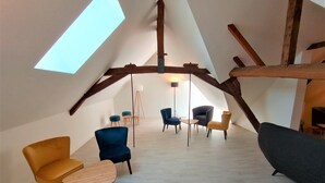 A spacious attic room with a sloped ceiling, exposed wooden beams, and a skylight. There are several chairs and a small table, with a floor lamp and a standing lamp providing lighting.