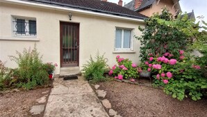 A two-story house with a tiled roof, a front door with a metal gate, and a small window with a curtain. There is a garden with pink flowers and a pathway leading to the door. - Le Florin d'Or - Bords de Creuse (Saint-Gaultier)