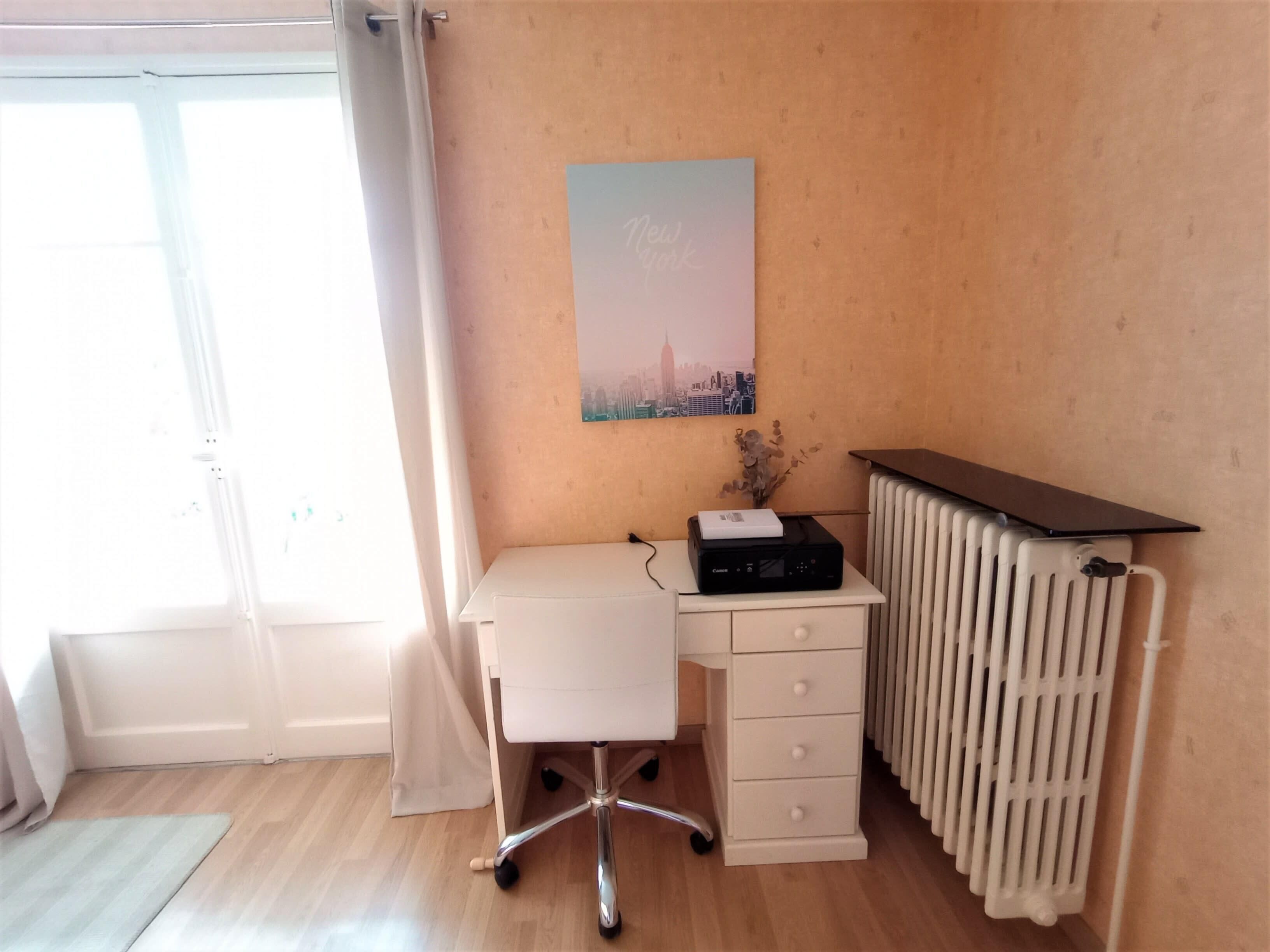 Et værelse med et hvidt skrivebord, en radiator og et bybillede på væggen.