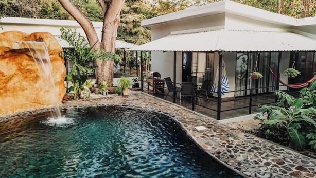 A pool with a waterfall, surrounded by stone walls and greenery, with a covered seating area and a hammock. - Villa Milu, Pool, Avellanas (Tamarindo)