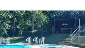 A swimming pool with white chairs around it, surrounded by greenery and a covered area in the background.