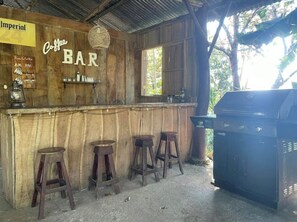 Un bar rustico con pareti in legno e un'insegna che recita 'Coffee BAR'. Tre sgabelli e una griglia si trovano sulla destra.