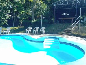 Une piscine entourée de chaises blanches, de verdure et d’un espace couvert en arrière-plan.