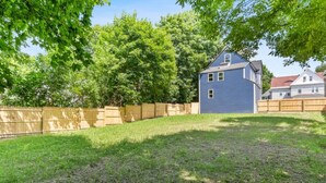 A backyard with a wooden fence, a grassy lawn, and a blue house with a gabled roof. - Fully Furnished 1st Floor Retreat, Comfort Awaits (Lewiston)