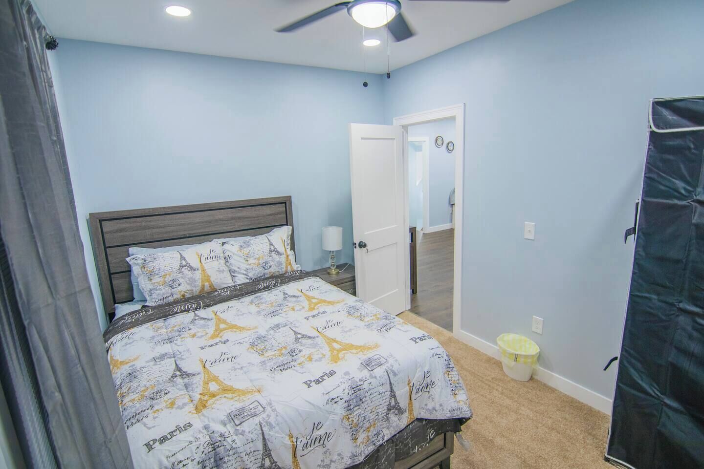 Un dormitorio con una cama, un ventilador de techo, una puerta y un pequeño cubo de basura.
