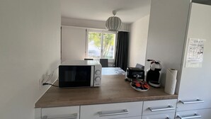 Kompakti keittiö, jossa on mikroaaltouuni, leivänpaahdin ja kahvinkeitin puupintaisella työtasolla. Sen alla on valkoiset kaapit ja ikkunassa on verhot.