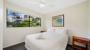 A hotel room with a large bed, a ceiling fan, a window with a view of a building and greenery, and a framed picture on the wall. - Unit 10 Marcoola Beach Resort (Marcoola)