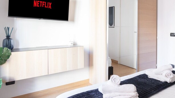 Moderni hotellihuone, jossa on litteänäyttöinen televisio, jossa näkyy Netflixin logo, puinen meikkipöytä peilillä sekä siististi pedattu sänky pyyhkeineen.