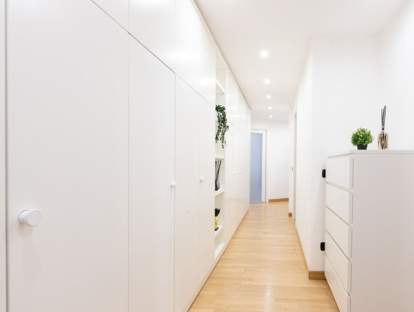 A narrow hallway with white cabinets and shelves, a wooden floor, and a door at the end.