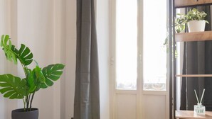 Huoneessa on ruukussa oleva monstera-kasvi, harmaa verho, puulattia, valkoinen ovi ja hylly, jossa on ruukussa oleva kasvi ja laatikko, jonka päällä on merkintä ”BIS”.