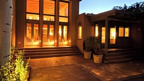 A modern house with a large glass window, a brick pathway, and a tree. - Casa Serenata - Gated Pueblo Retreat Near Opera, Views + Office - Monthly (Santa Fe)