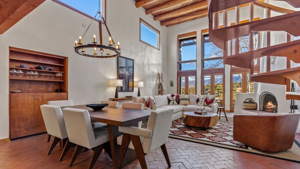 A spacious living area with a wooden dining table, comfortable seating, a fireplace, and large windows offering a view of the outdoors. - Casa Serenata - Gated Pueblo Retreat Near Opera, Views + Office - Monthly (Santa Fe)