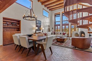 A spacious living area with a wooden dining table, comfortable seating, a fireplace, and large windows offering a view of the outdoors. - Casa Serenata - Gated Pueblo Retreat Near Opera, Views + Office - Monthly (Santa Fe)