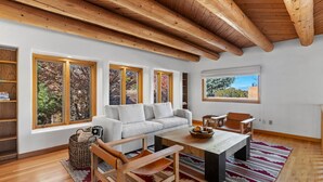 A modern living room with a white sofa, wooden coffee table, and a view of trees through large windows. - Casa Serenata - Gated Pueblo Retreat Near Opera, Views + Office - Monthly (Santa Fe)