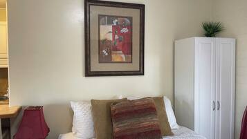 Un dormitorio ordenado con cama, almohadas, un cuadro enmarcado, un armario y un escritorio con lámpara.