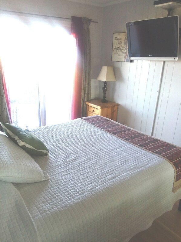 A neatly made bed with pillows, a bedside lamp, a flat-screen TV mounted on the wall, and a window with curtains.